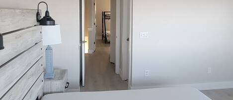 A bedroom with a bed, a nightstand, a lamp, and a ceiling fan. There is a hallway leading to another room.
