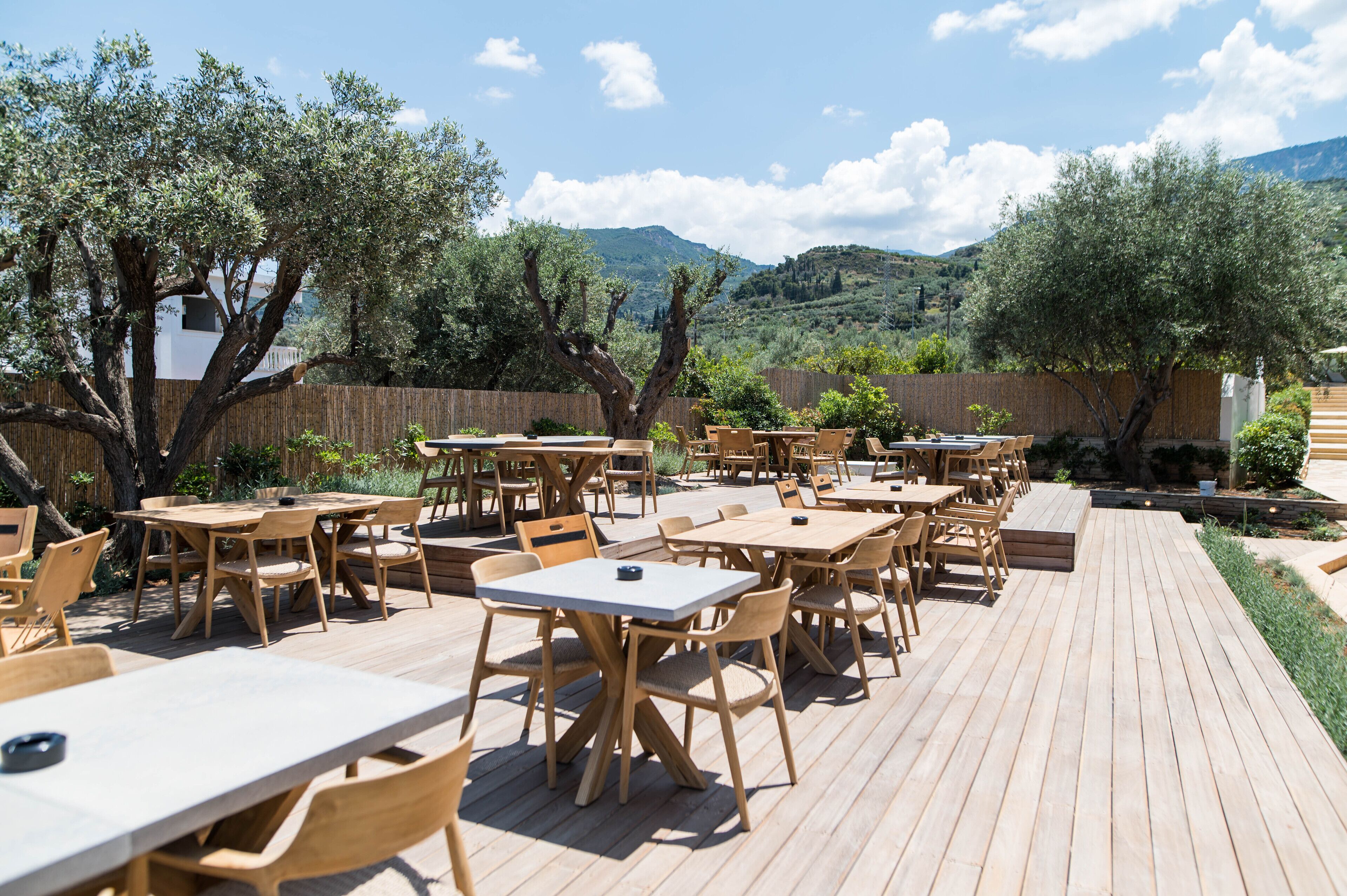 An outdoor patio with wooden tables and chairs, surrounded by trees and a view of hills.
