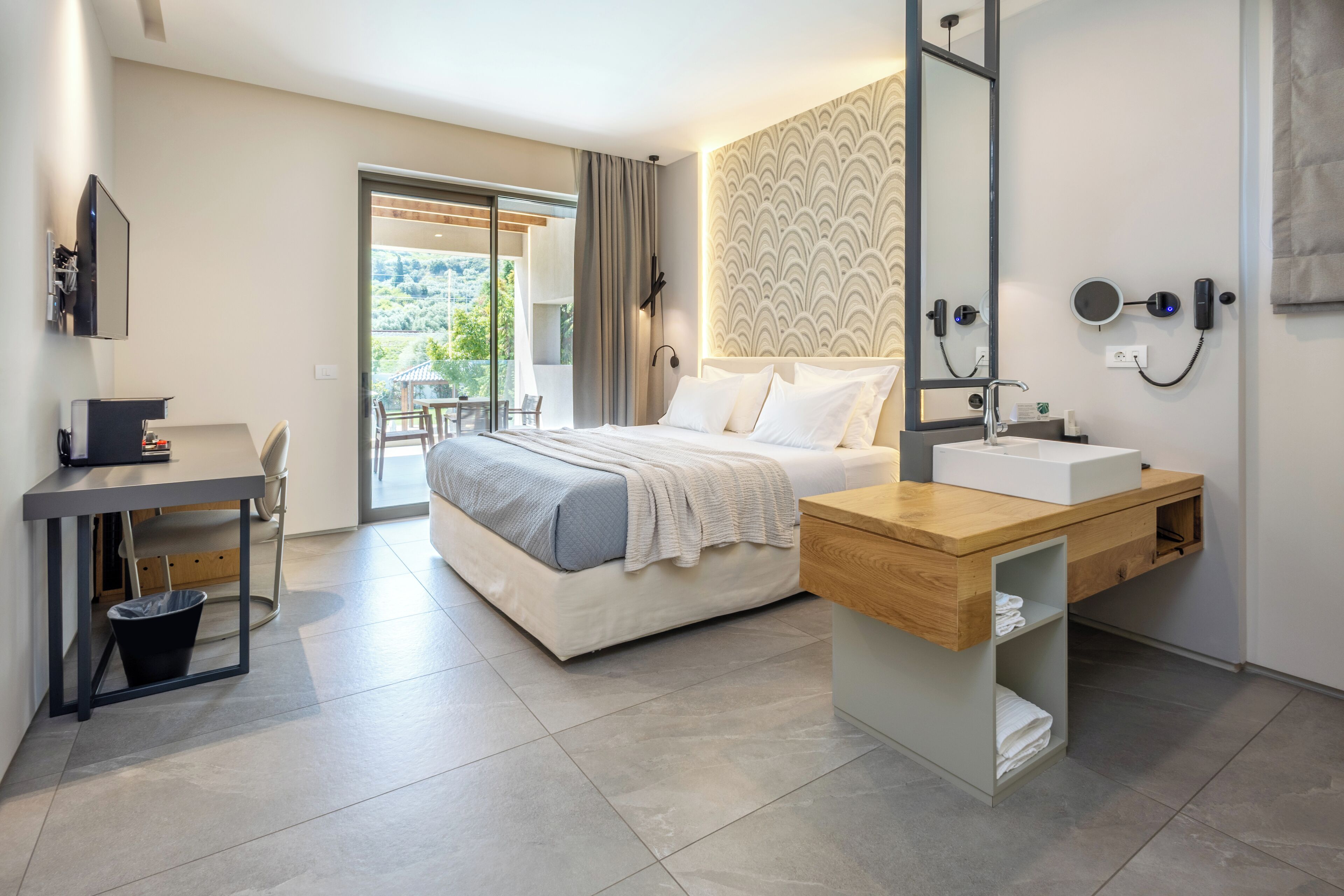 A modern hotel room with a bed, a desk, a sink, and a balcony with a view of greenery.