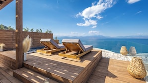 Eine Holzdielen-Terrasse mit zwei Liegestühlen und einer Kerze, mit Blick auf das klare, blaue Meer und ferne Berge.