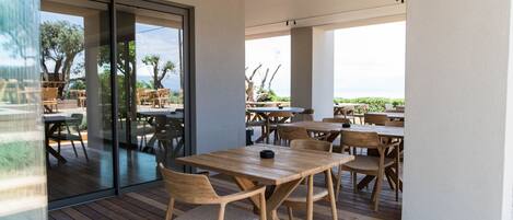 An outdoor dining area with wooden tables and chairs, large windows, and a view of the outdoors.