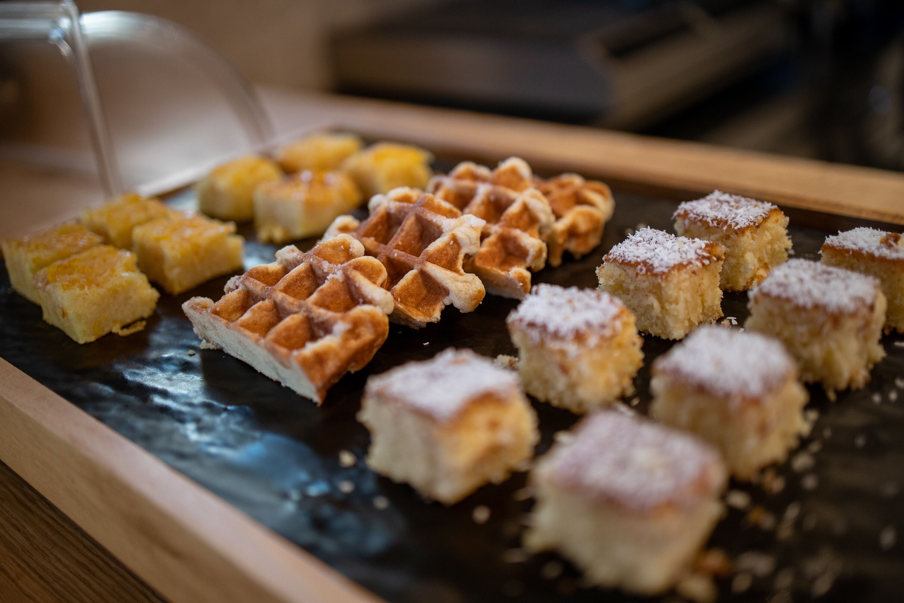 A tray of assorted pastries, including waffles and cake squares, on a wooden surface.
