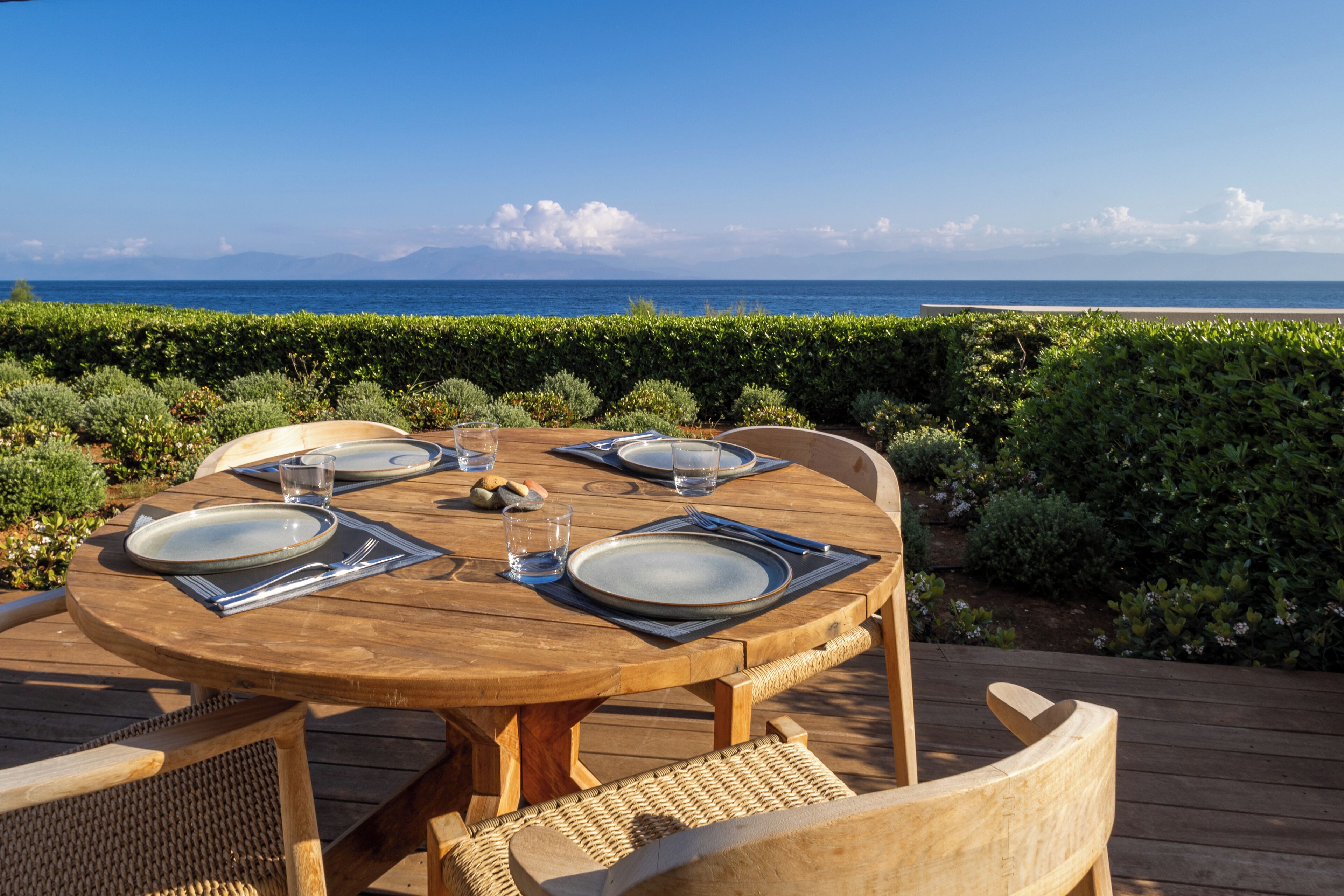 Une table en bois, dressée pour quatre personnes, avec des assiettes, des couverts et des verres, donnant sur une plage et l’océan.