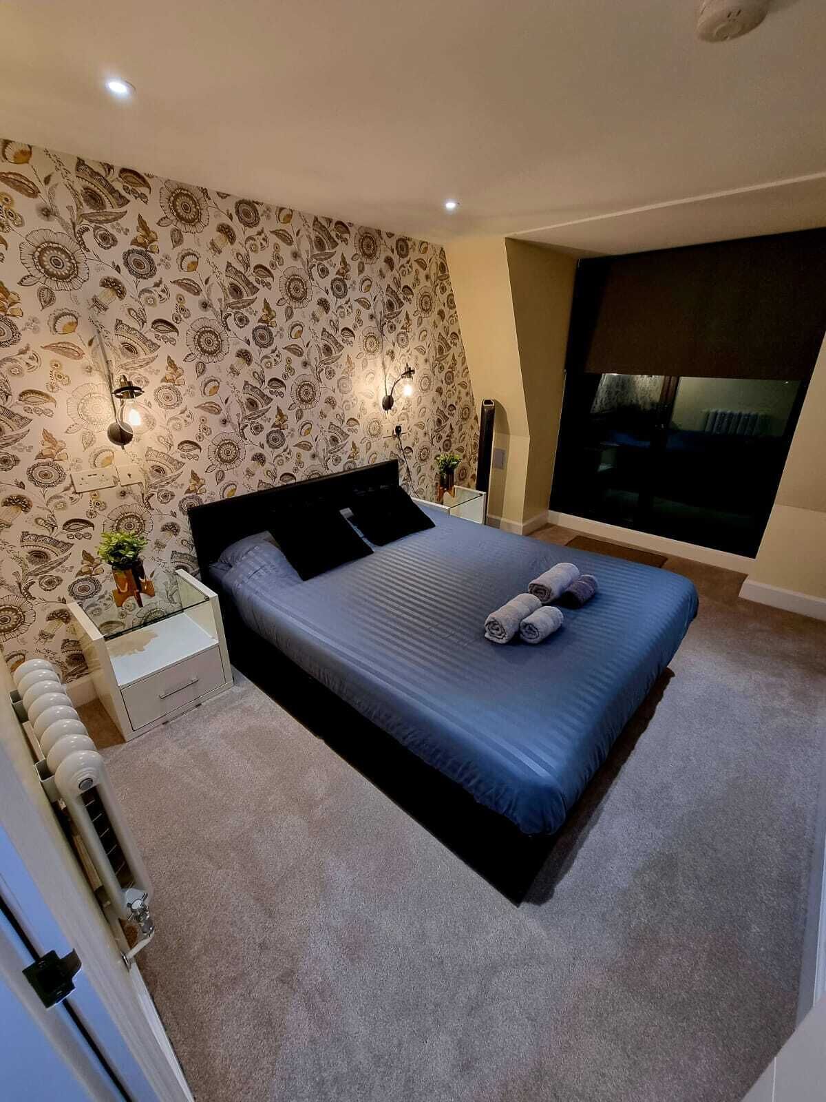A modern bedroom with a large bed, patterned wallpaper, bedside lamps, and a view of the city at night.