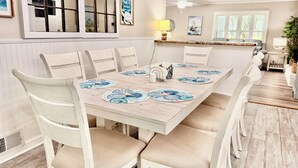 A dining area with a white table set with blue and white plates, surrounded by white chairs with beige cushions. - Hidden Treasure: Close to Beach with Rooftop Deck, Dog Friendly, Fenced Yard (Oak Island)
