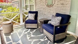 A covered porch with two blue armchairs, a wicker table, and a brick wall with a turtle logo. - Hidden Treasure: Close to Beach with Rooftop Deck, Dog Friendly, Fenced Yard (Oak Island)