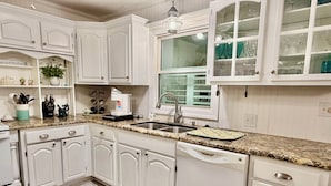 A kitchen with white cabinetry, a granite countertop, a coffee maker, and a window with curtains. - Hidden Treasure: Close to Beach with Rooftop Deck, Dog Friendly, Fenced Yard (Oak Island)