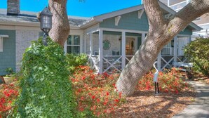 A two-story building with a porch, surrounded by trees and red flowers. - Hidden Treasure: Close to Beach with Rooftop Deck, Dog Friendly, Fenced Yard (Oak Island)
