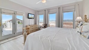 Una camera da letto con un letto grande, un ventilatore a soffitto, una TV e vista sulla spiaggia attraverso le porte scorrevoli in vetro.