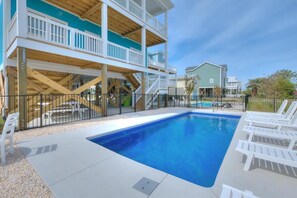 A swimming pool with a wooden deck, white lounge chairs, and a multi-level building with balconies.