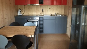 Een kleine keuken met rode kastjes, een spoelbak, een fornuis en een koelkast. Er staat een eettafel met stoelen.