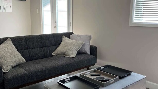 A modern living room with a grey sofa, a grey ottoman, and a window with blinds. - Lake Simcoe Beach Oasis with Backyard and Firepit (Willow Beach)