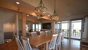 A spacious dining area with a long wooden table and white chairs, large windows, and a view of the outdoors. - Primehook Beach Bayfront Beauty (Milford)
