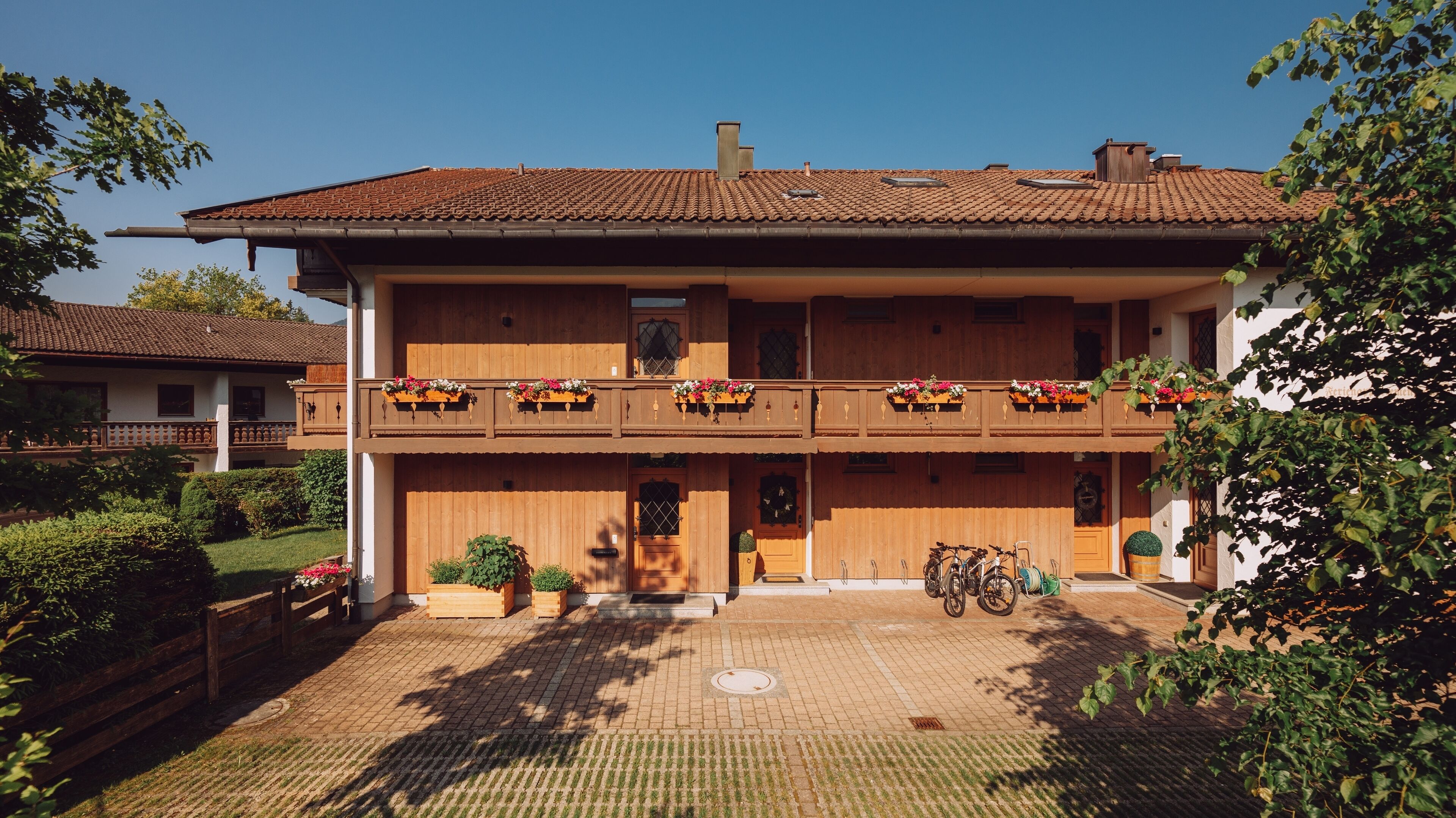 A two-story wooden house with a tiled roof, a balcony with flower boxes, and a paved driveway.