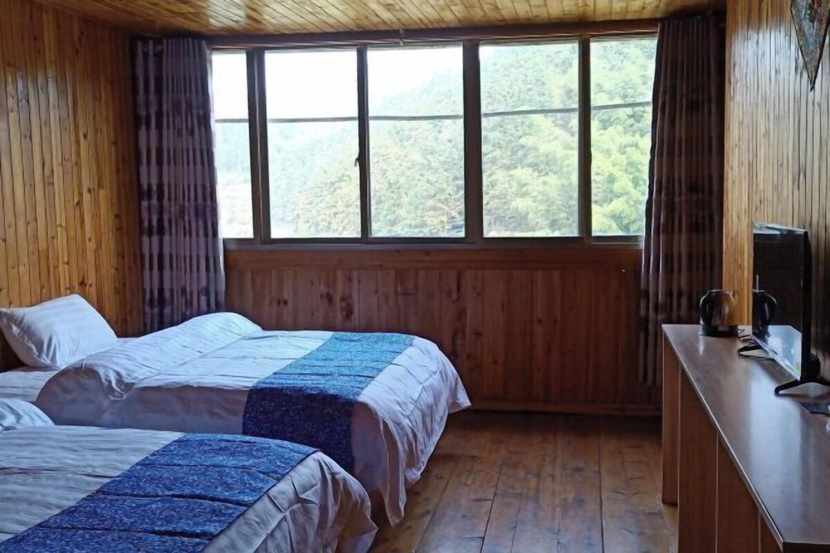 A wooden room with two beds, a wooden desk, and a window with a forest view.
