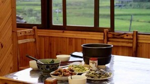 Un intérieur en bois avec une table à manger dressée pour un repas, une fenêtre donnant sur un paysage verdoyant et un grand pot noir posé sur la table.