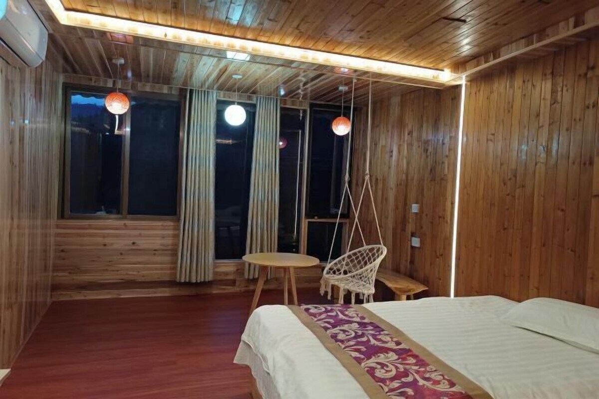 A wooden room with a bed, a hanging chair, a small round table, and a window with curtains.