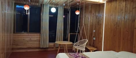 A wooden room with a bed, a hanging chair, a small round table, and a window with curtains.
