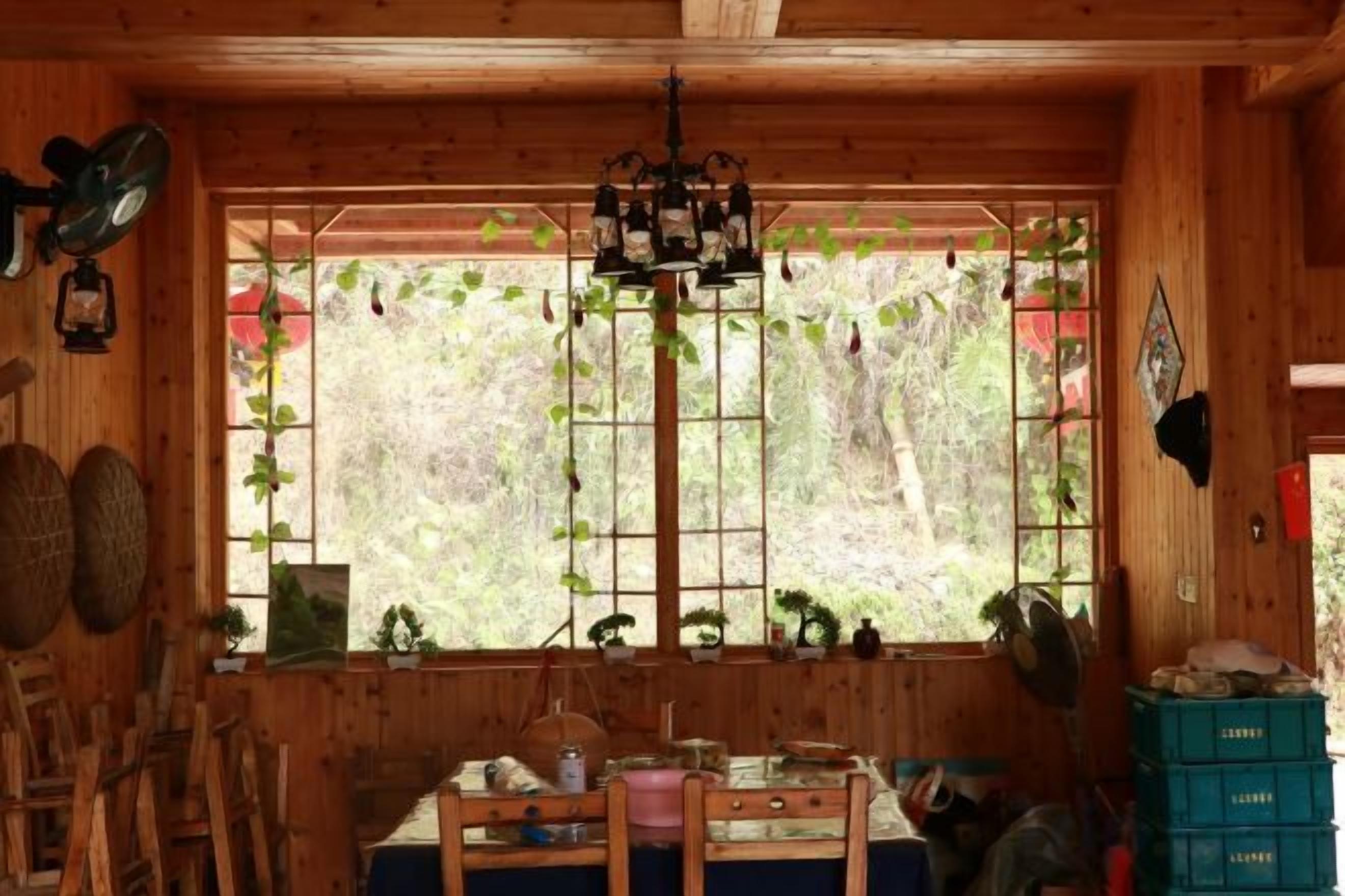 Innenansicht einer Holzhütte mit einem Esstisch, Stühlen, einer Chandelierleuchtre und einem Fenster mit Pflanzen.