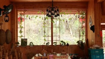 Innenansicht einer Holzhütte mit einem Esstisch, Stühlen, einer Chandelierleuchtre und einem Fenster mit Pflanzen.