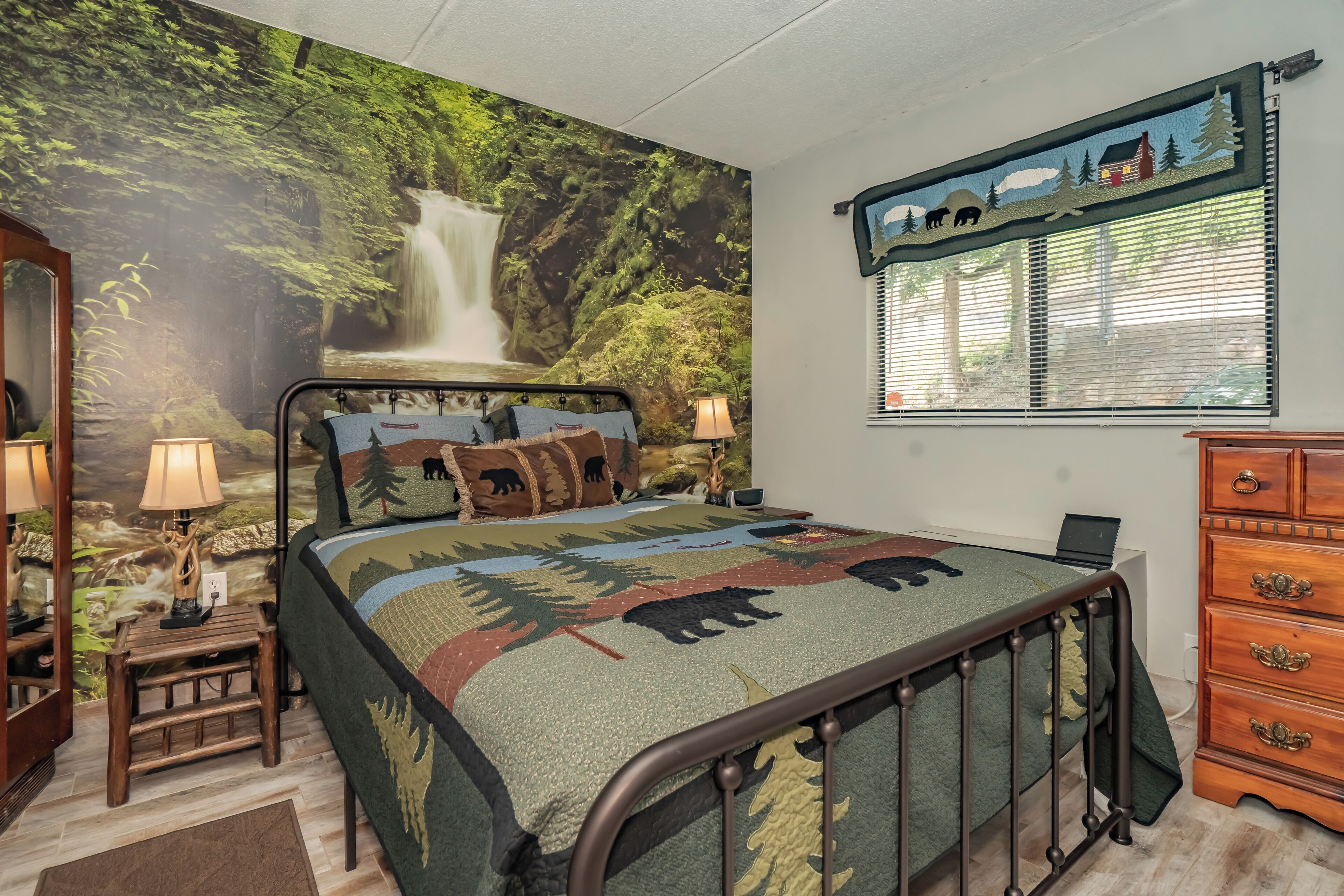 Une chambre à coucher avec une fresque murale sur le thème de la forêt, un lit recouvert d’une courtepointe à motifs d’ours, une table de chevet avec une lampe, une commode et une fenêtre avec des stores.