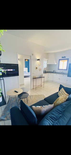A modern living room with a blue sofa, a small round table, and a potted plant. There is a kitchen area with white cabinets and a window. - Apartments in private residence with sea view, quiet and close to shops and beaches. (Solenzara)