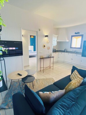 A modern living room with a blue sofa, a small round table, and a potted plant. There is a kitchen area with white cabinets and a window.