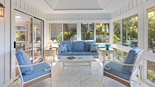 Una habitación soleada con un sofá azul, dos sillones blancos, una mesa de centro y ventanas amplias con vistas al verdor exterior.