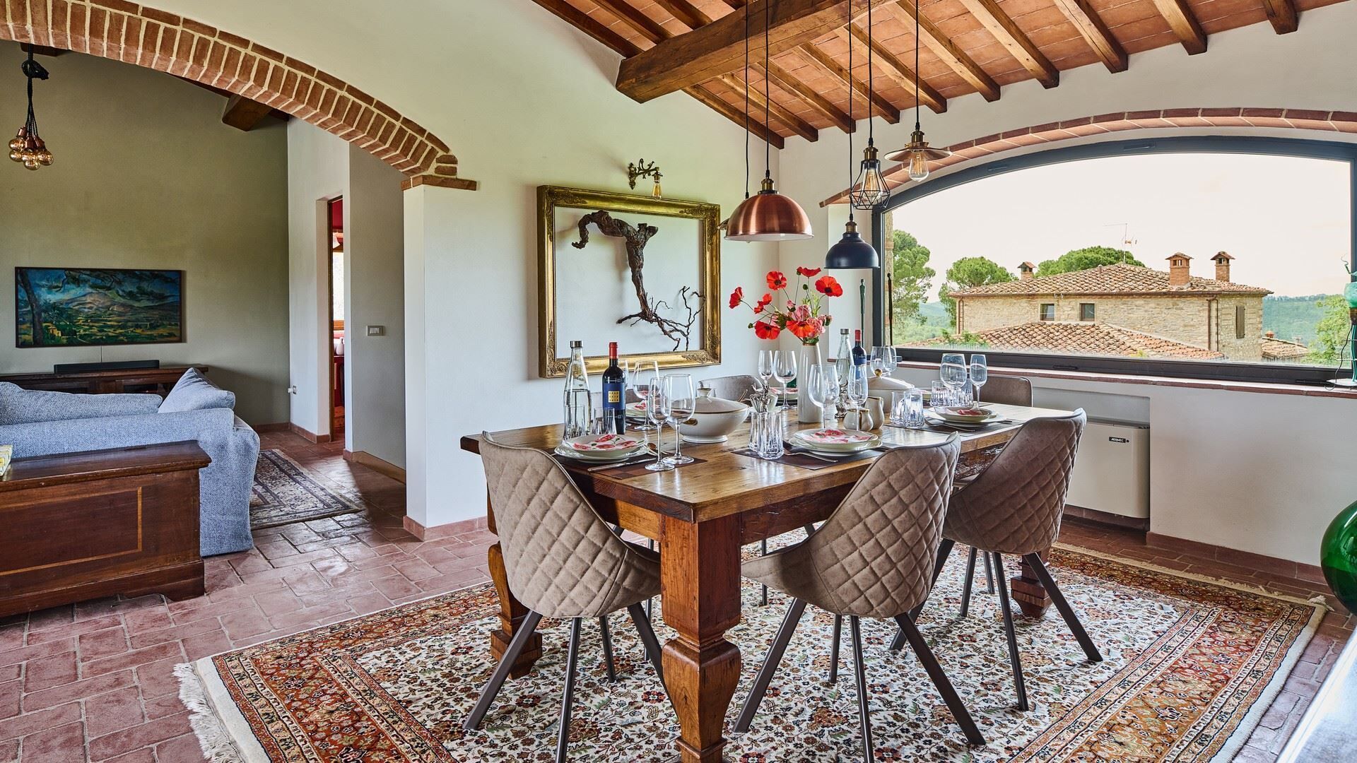 A dining area with a wooden table set for six, surrounded by quilted chairs, a decorative mirror, and a view of the countryside through a large window.