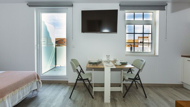 Una habitación pequeña y luminosa con una cama, una mesa con sillas, un televisor y dos ventanas.