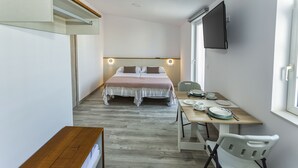 Habitación compacta con cama, escritorio y silla. Hay un televisor montado en la pared y una ventana que proporciona luz natural.
