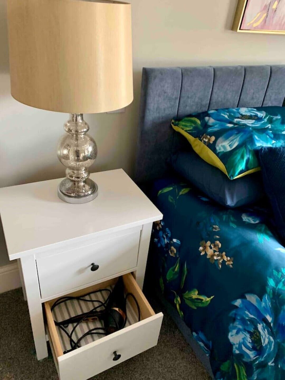 A bedroom with a white nightstand, a drawer with cables, a lamp, and a bed with blue floral bedding.