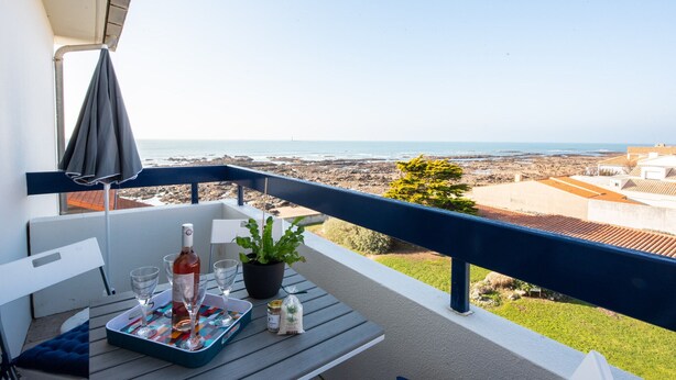 A balcony with a table set for two, a bottle of wine, and a potted plant.