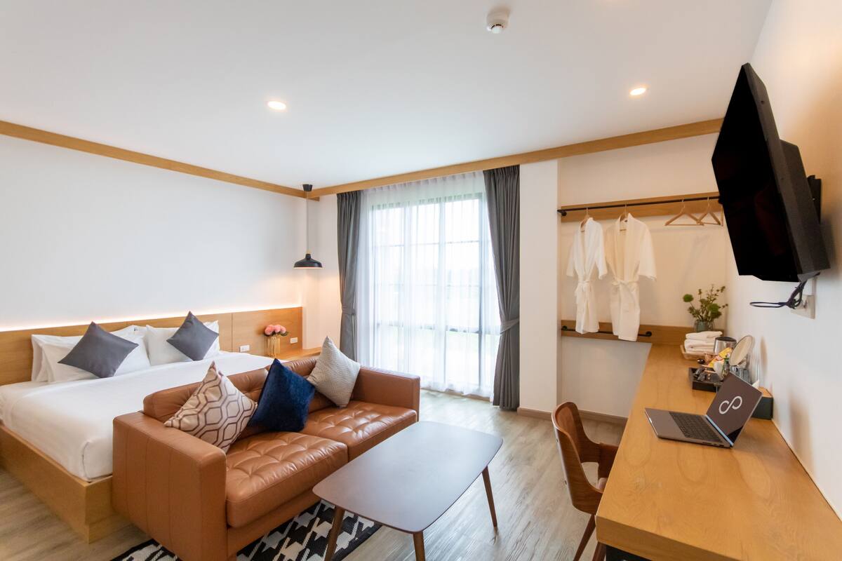 A hotel room with a bed, a sofa, a desk with a laptop, a TV, and a window with curtains.