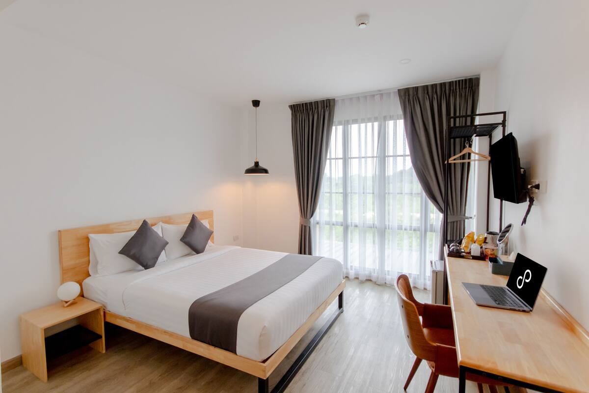 A hotel room with a bed, a desk with a laptop, a chair, a lamp, and a coat rack.