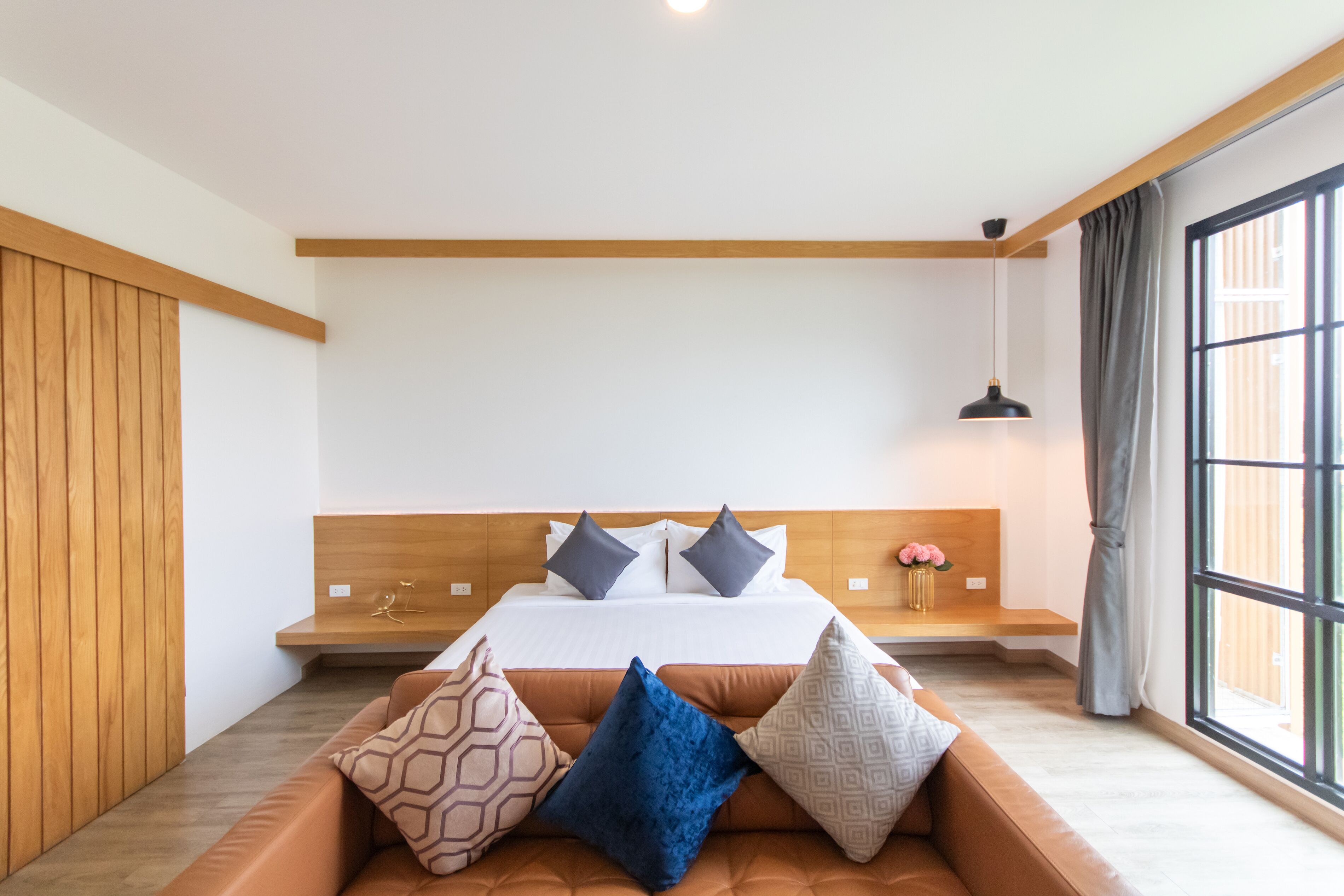 Un dormitorio moderno con cabecera de madera, una cama grande con muchas almohadas, un banco al pie de la cama y un ventanal con cortinas.