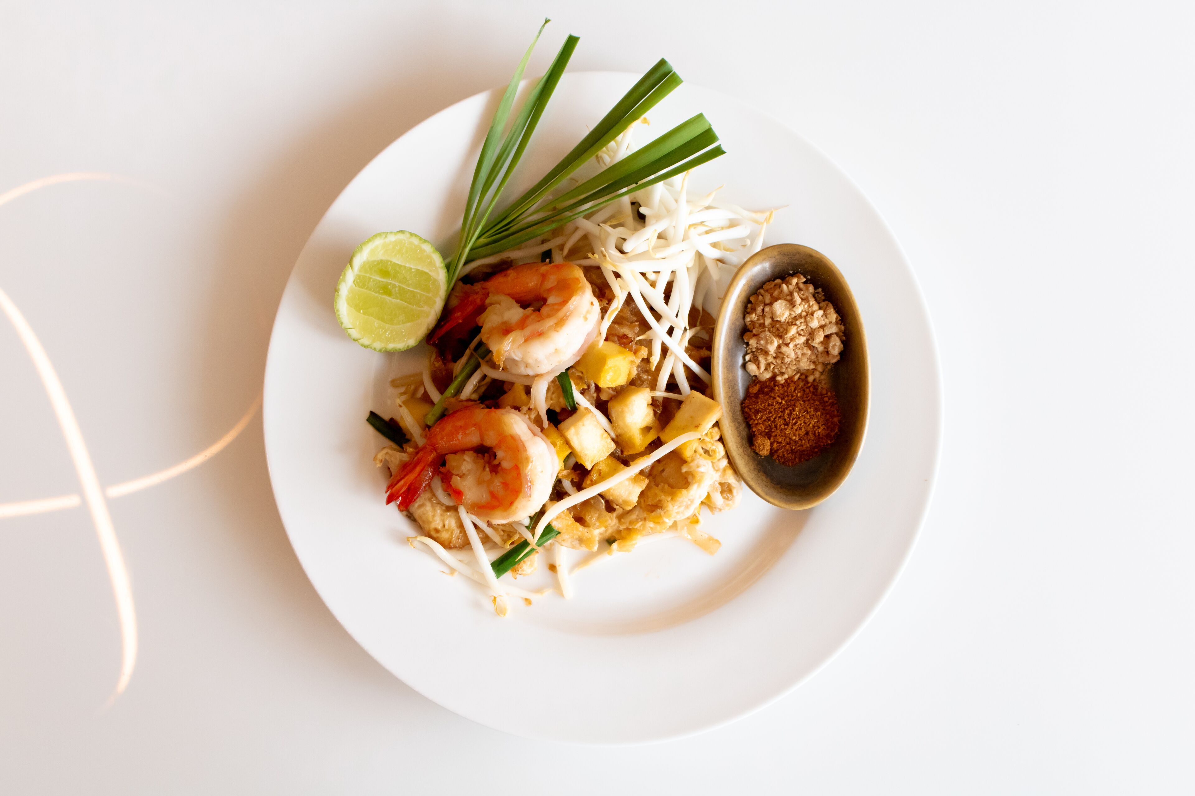 A plate of Pad Thai with shrimp, bean sprouts, lime, and a small bowl of sauce.