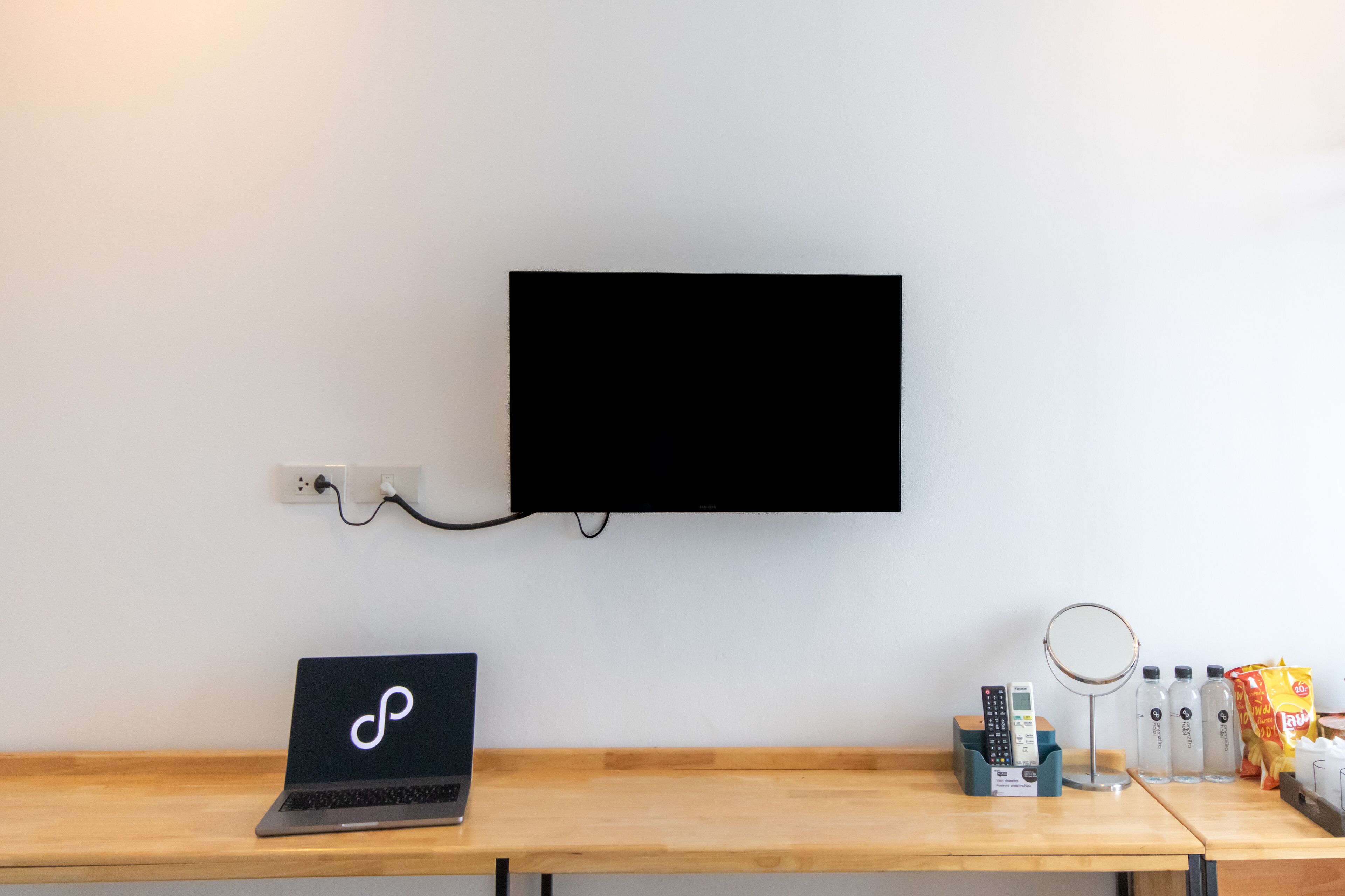 A laptop on a wooden desk, a mounted TV, a remote control, a magnifying glass, and a snack bag.