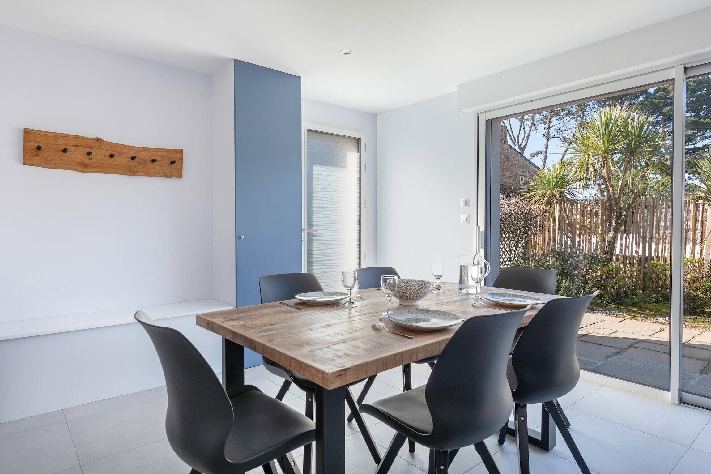Uma área de jantar moderna com mesa de madeira posta para quatro pessoas, cadeiras pretas e vista para o jardim exterior através de grandes portas de vidro.
