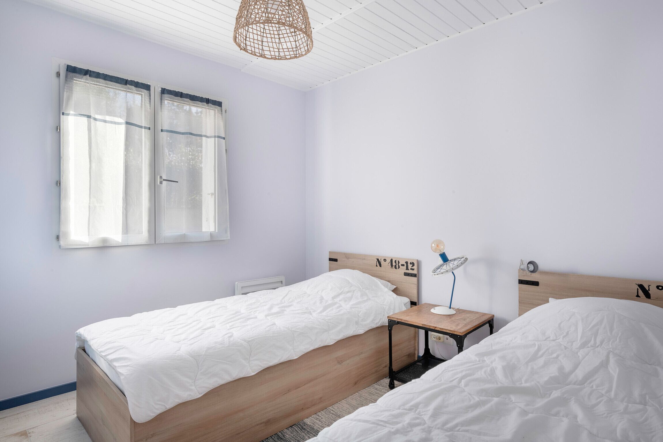 Un dormitorio con dos camas, una mesita de noche con lámpara, una ventana con cortinas y un ventilador de techo.