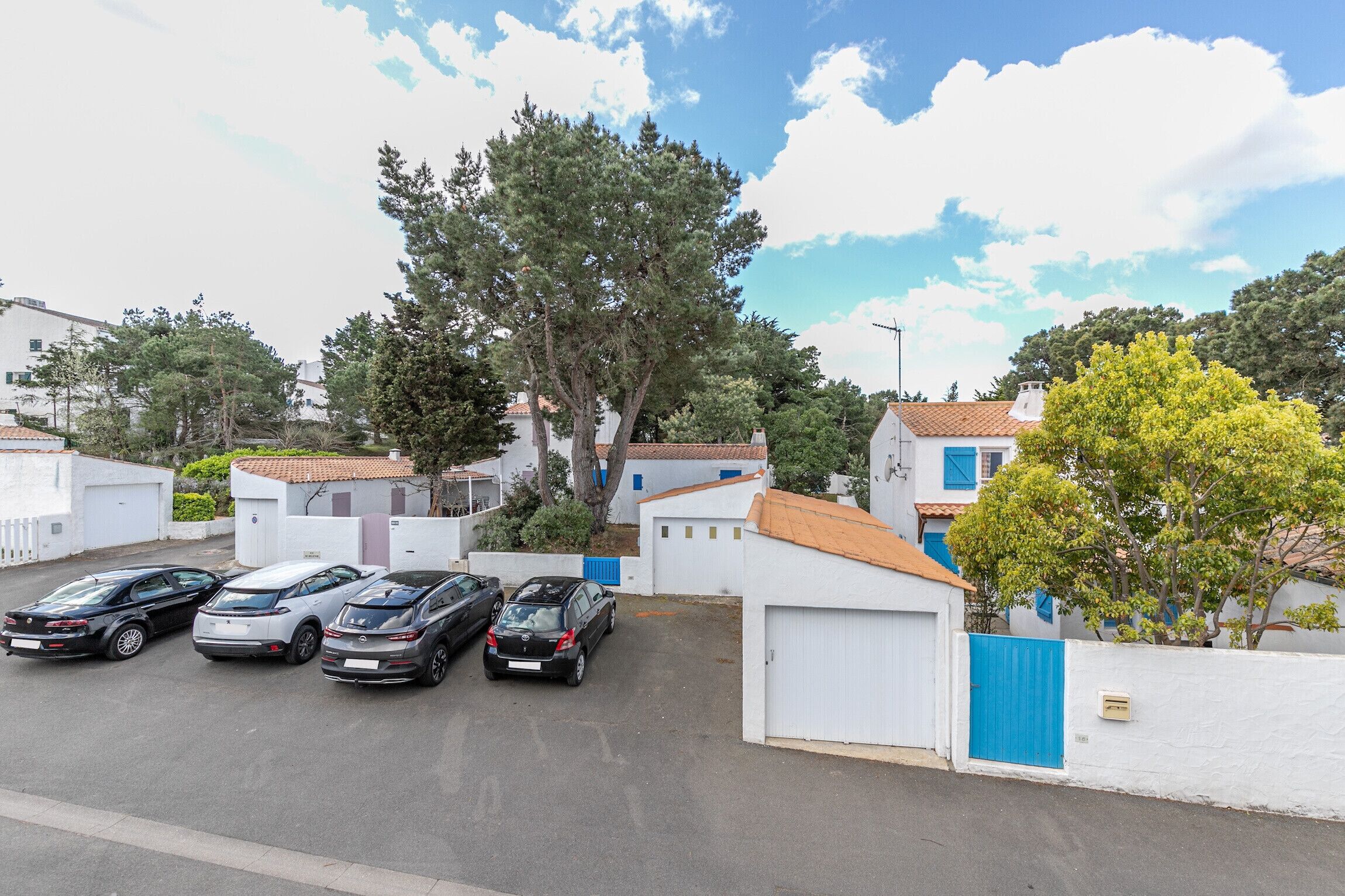 Un quartier résidentiel avec des maisons blanches, un parking pouvant accueillir cinq voitures et un grand arbre.