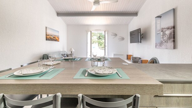 Un comedor moderno con una mesa larga dispuesta para cuatro personas, un ventilador de techo y un amplio ventanal que proporciona luz natural.