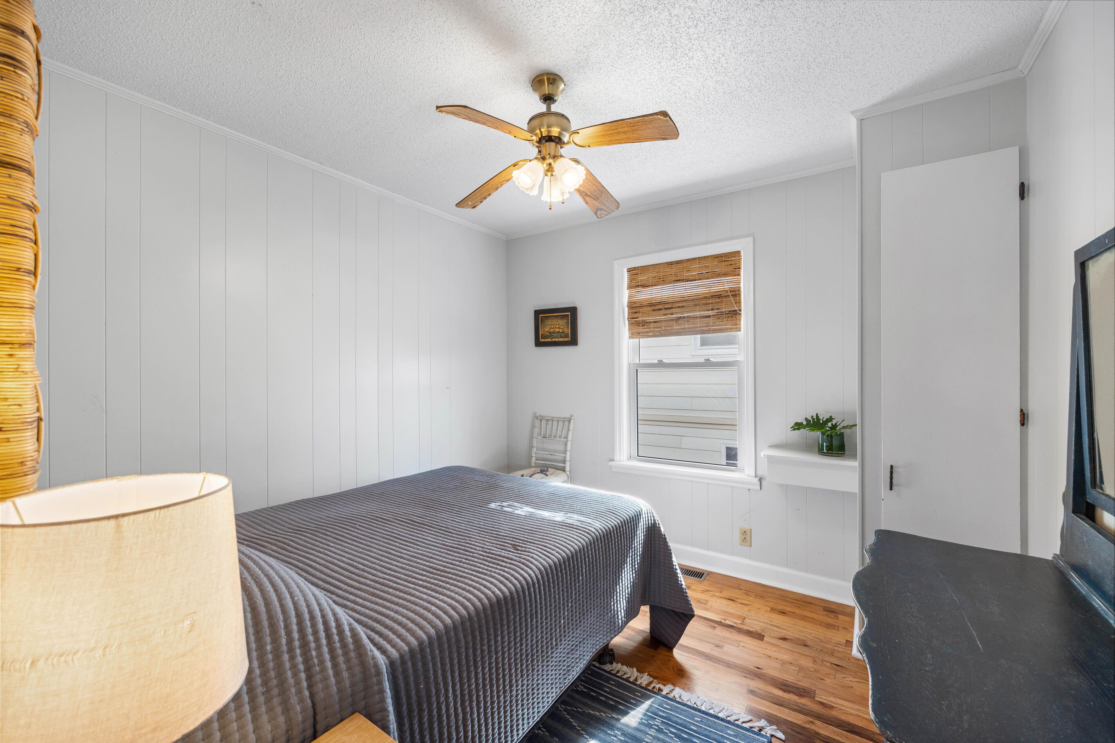 Une chambre à coucher avec un lit, un ventilateur de plafond, une fenêtre avec des stores, une petite plante et une chaise.