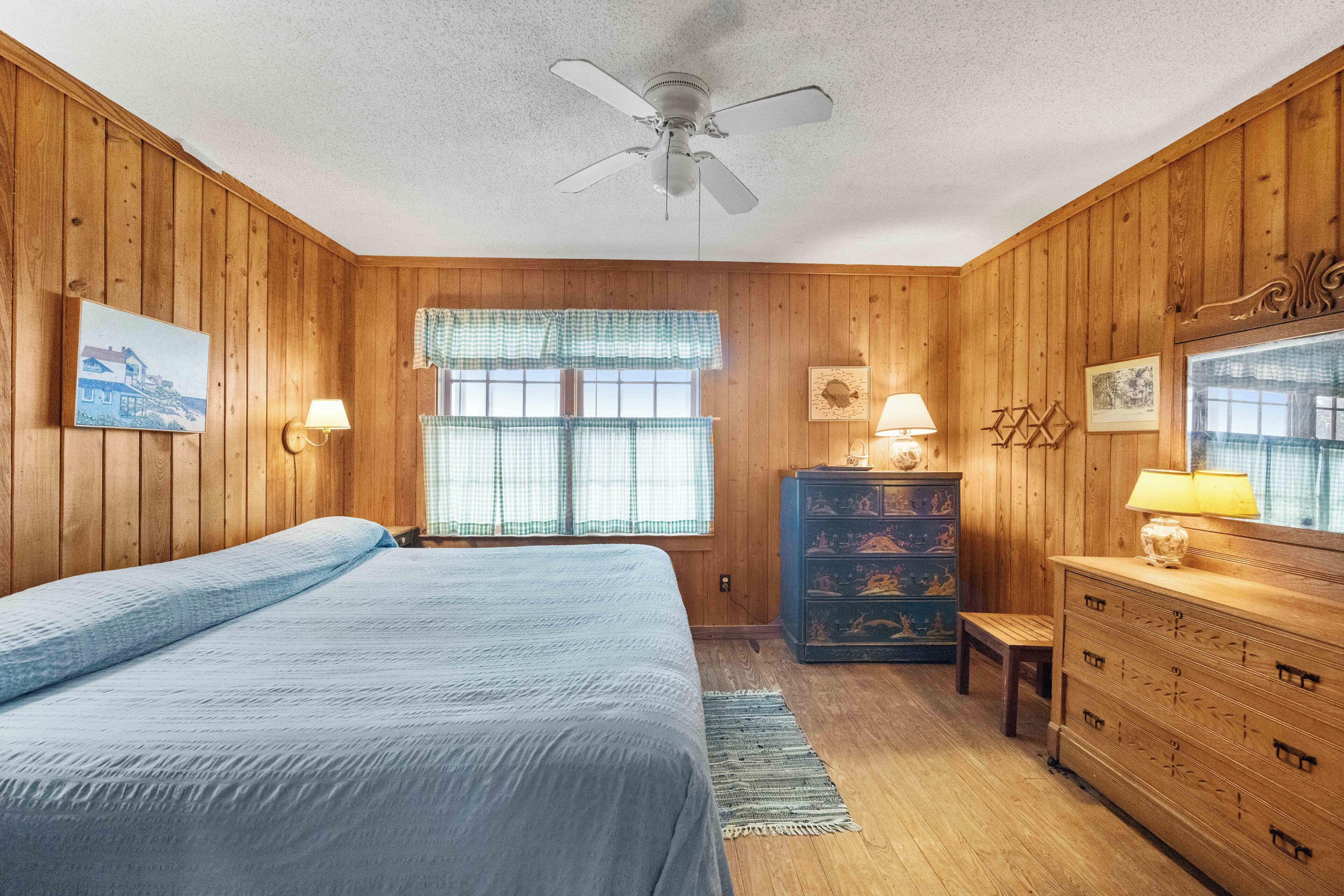 Une pièce aux murs en bois, avec un lit, une commode, un banc, une fenêtre avec des rideaux et un ventilateur de plafond.