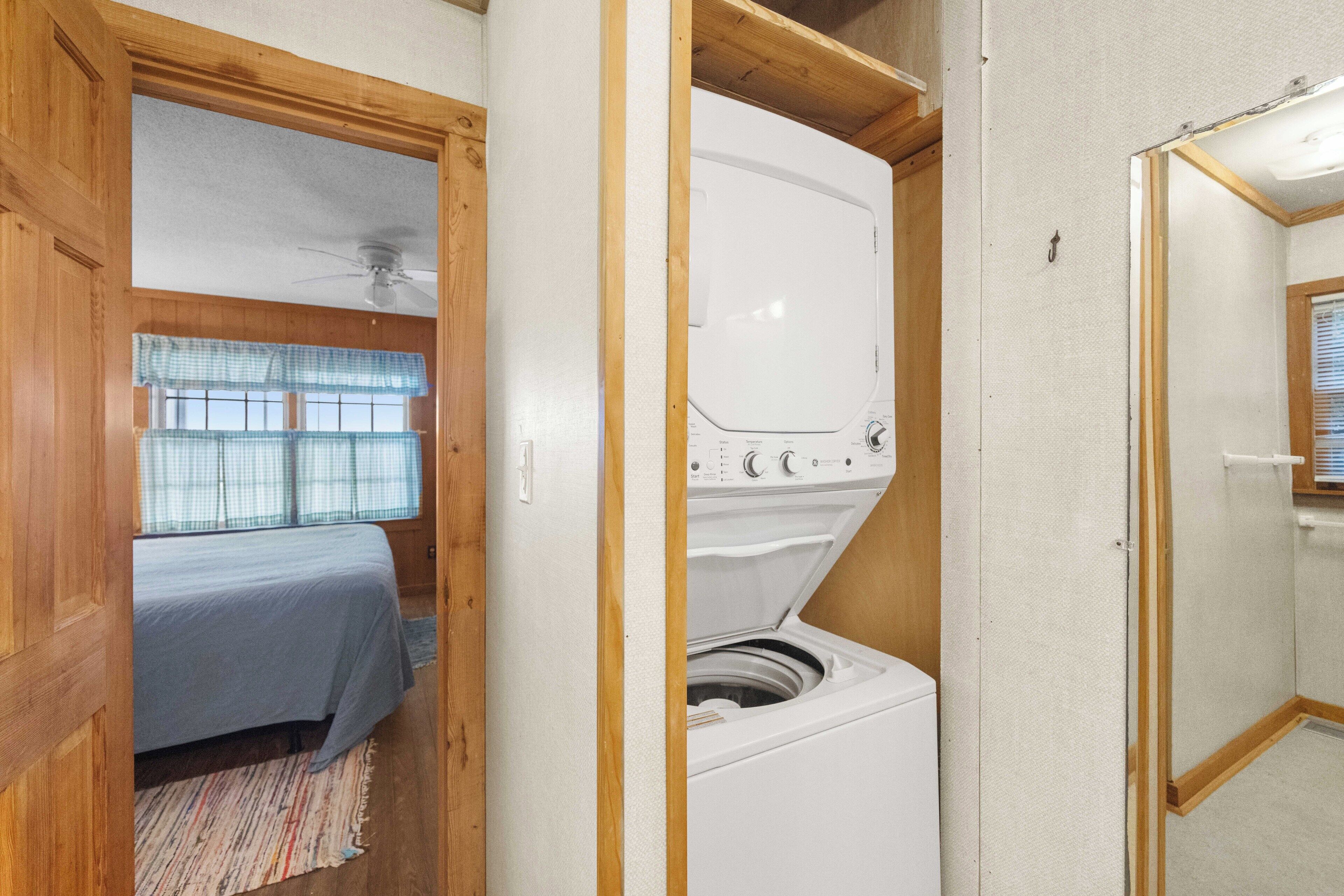 Une petite pièce comprenant un lit, un lave-linge et une petite fenêtre avec des stores.