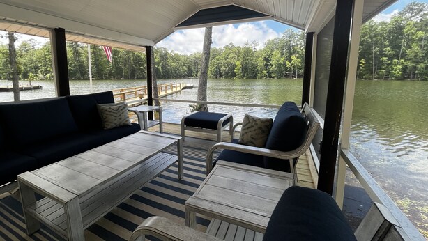 Un porche cubierto con un sofá, sillas y una mesa, con vista a un lago con un muelle y árboles.
