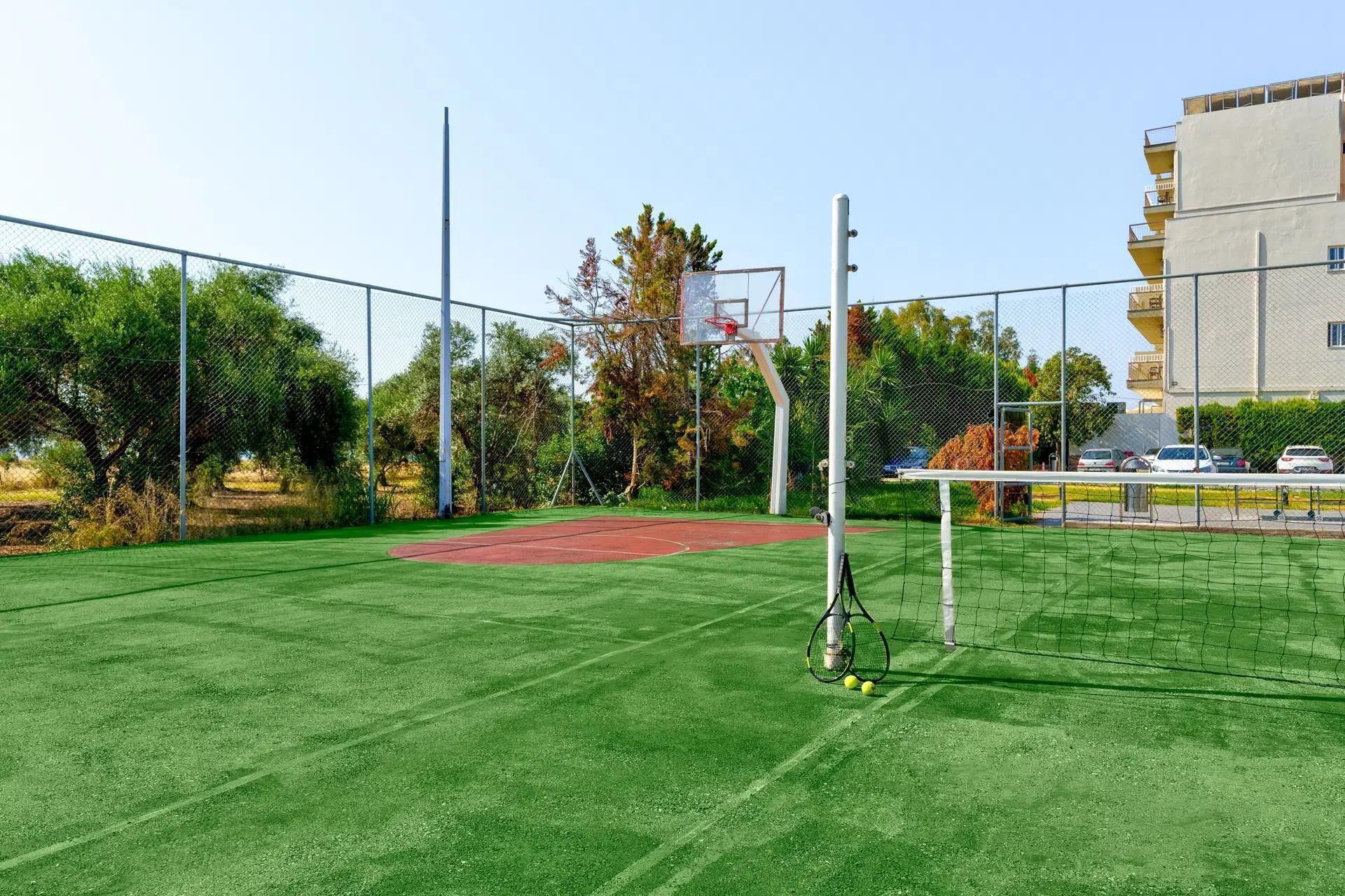 An outdoor tennis court with a basketball hoop, surrounded by a chain-link fence and trees.