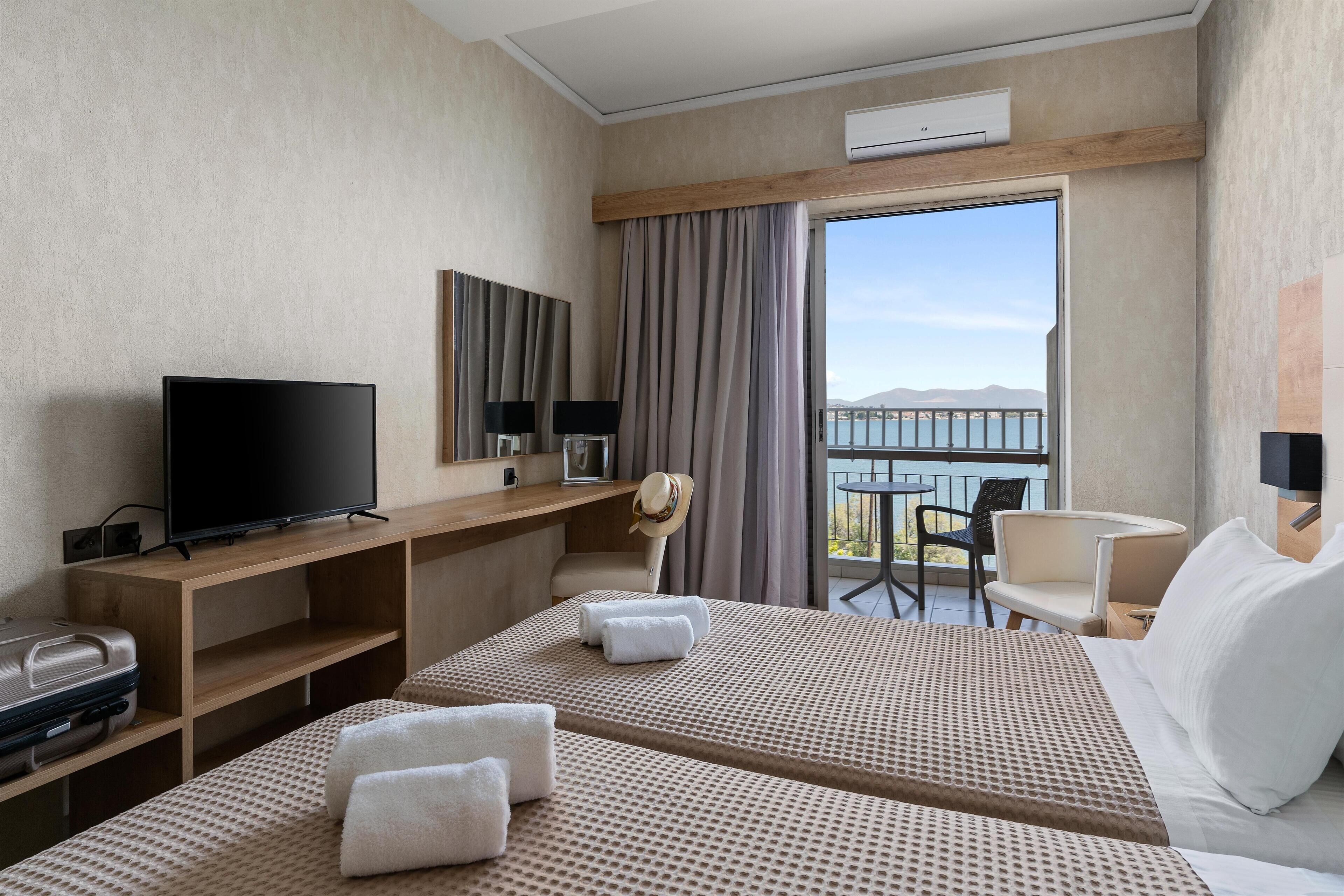 A hotel room with two beds, a TV, a desk, and a balcony with a view of the sea.