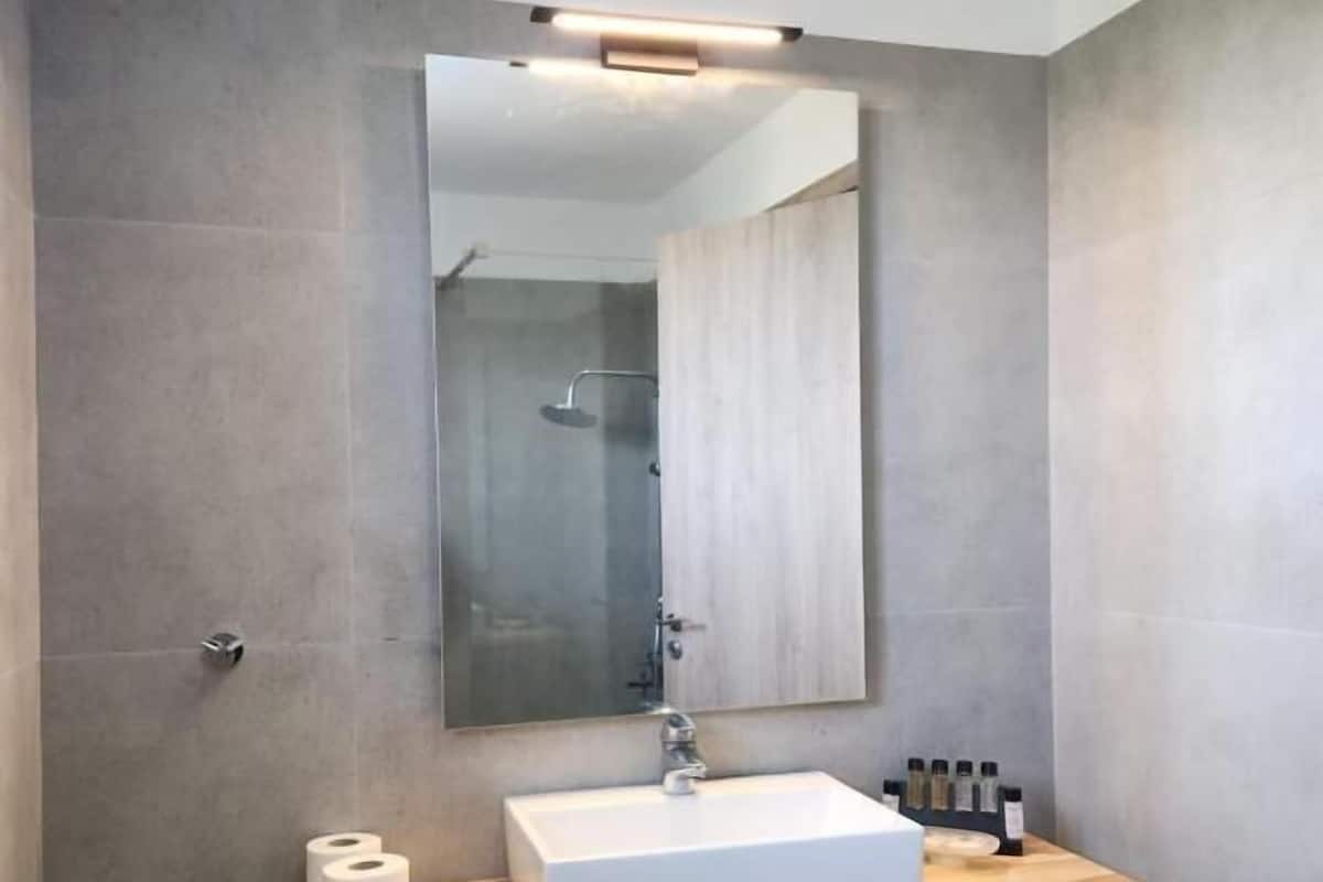 A modern bathroom with a large mirror, a white sink, and a wooden shelf with toiletries.