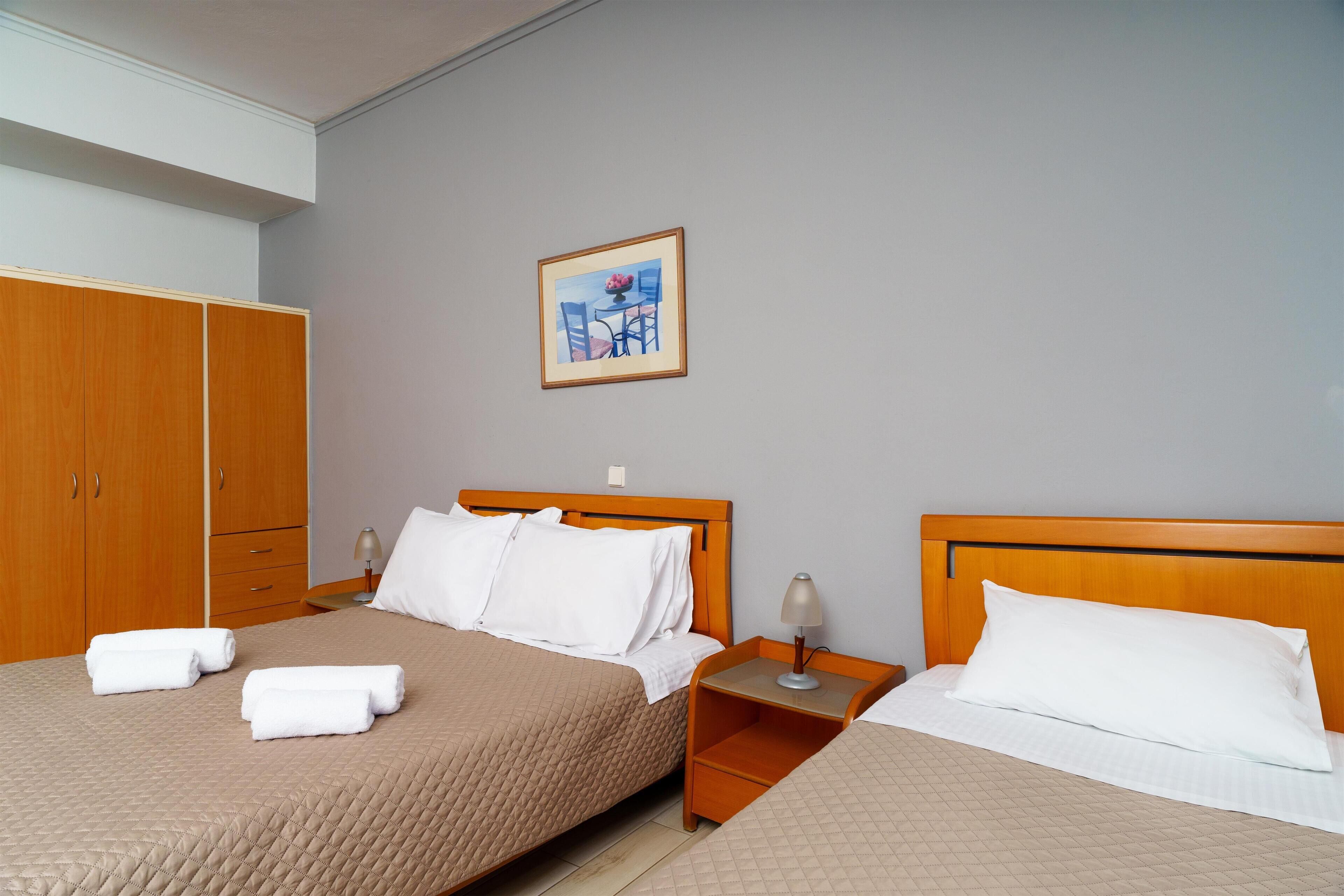 A hotel room with two beds, a wooden wardrobe, a nightstand with a lamp, and a framed picture on the wall.